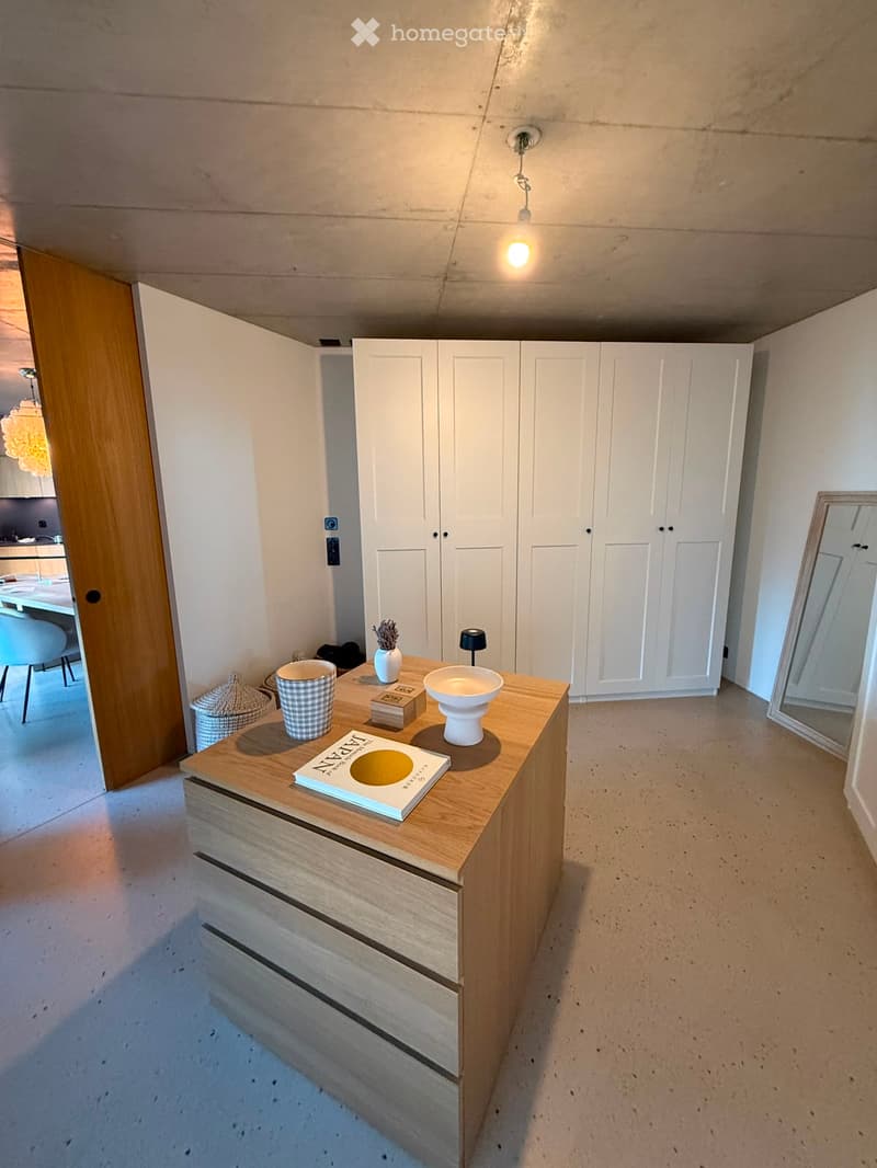 A modern, minimalist room with a wooden table holding a cup, bowl, book, and decorative items. White cabinetry and a large mirror are visible, with a view into an adjacent dining area.