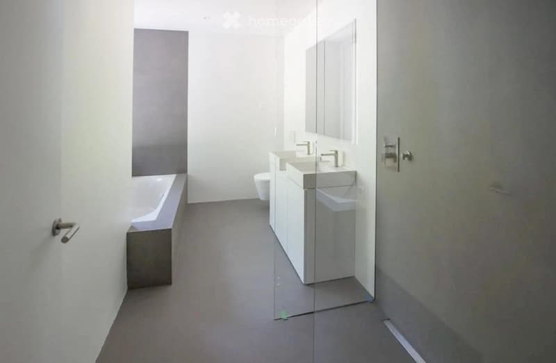 Modern bathroom with a bathtub, two sinks, a toilet, and a large mirror. The space features a glass partition and sleek, minimalist design.