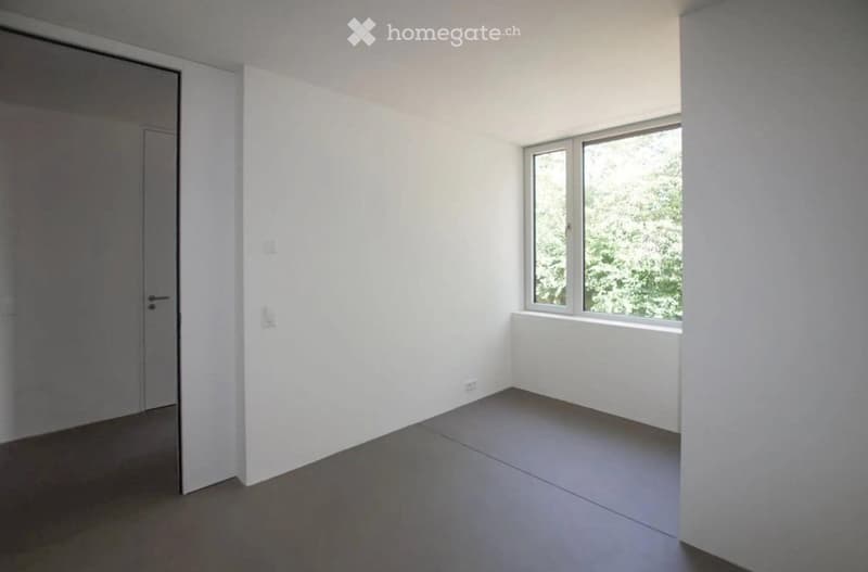 Empty room with white walls, grey floor, and a window with a view of greenery.