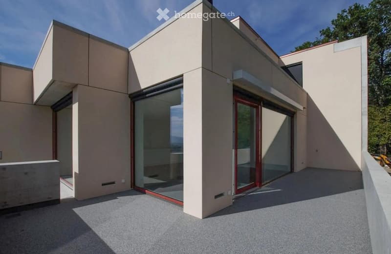 Modern two-story building with beige walls, large glass windows, and a raised ground floor. Features a balcony and a spacious outdoor area.