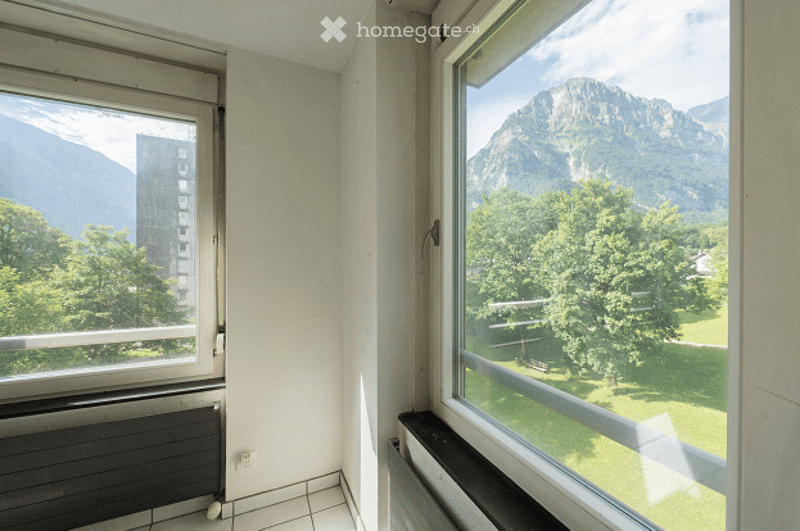 Empty room with large windows offering a scenic mountain view and a cityscape. The room has tiled flooring and radiators under the windows.