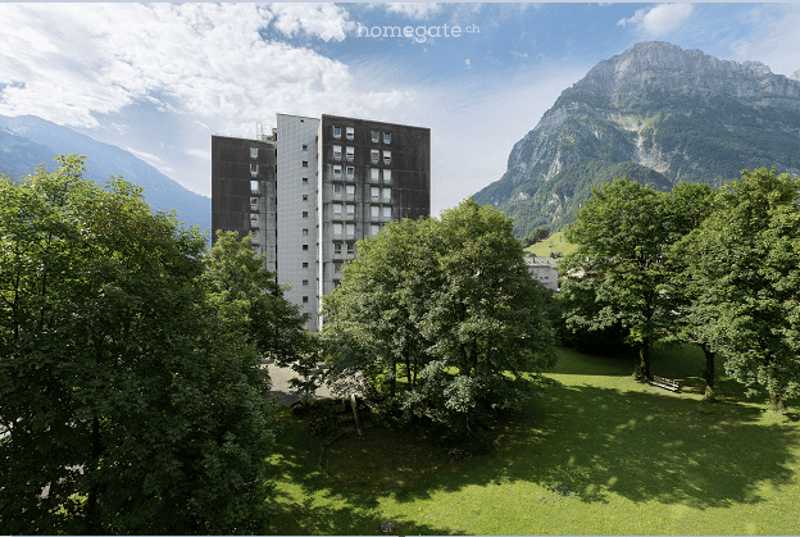A modern apartment building surrounded by lush green trees and a scenic mountain backdrop. The building has multiple floors with numerous windows and is situated in a serene, green landscape.