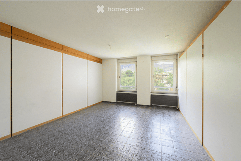 Completely empty room with grey tiled floor, white walls, and large windows with radiators underneath. No furniture or equipment present.