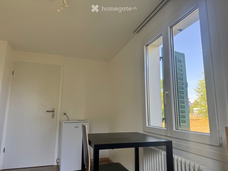 A small, furnished room with a table, chair, and a small refrigerator. The room has large windows with a view of the outside, a door, and radiators along the wall.