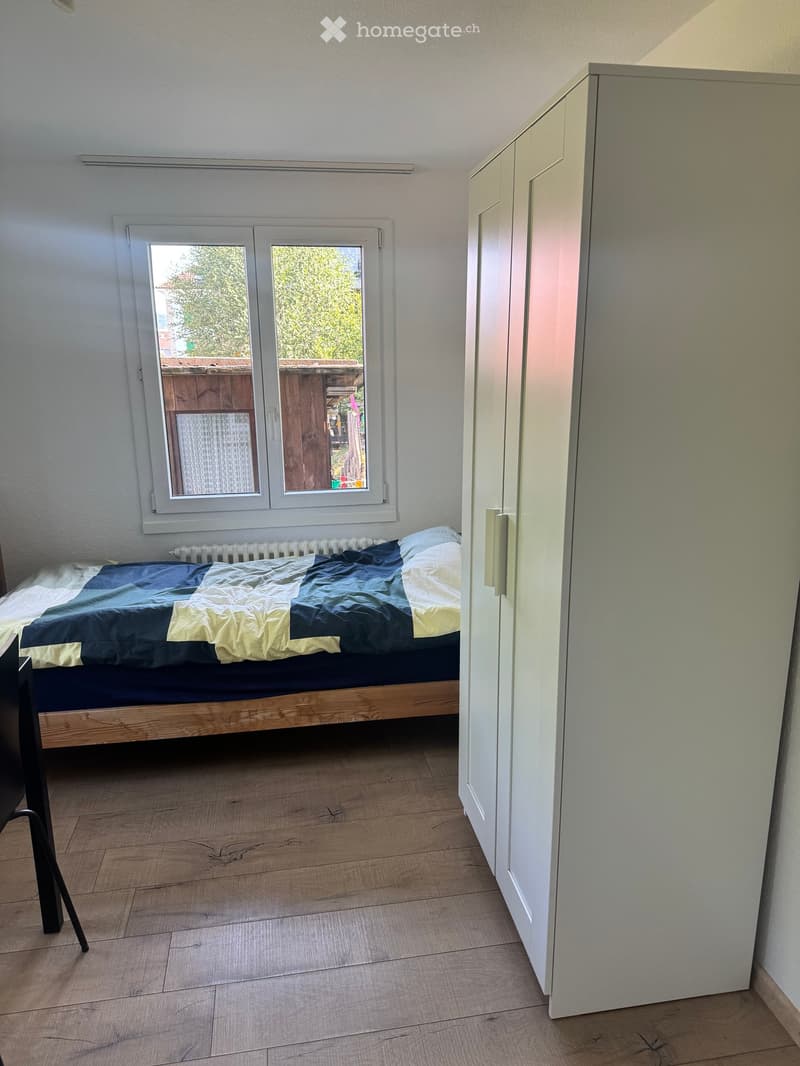 Bedroom with wooden floor, large window, bed with blue and yellow bedding, and white wardrobe.