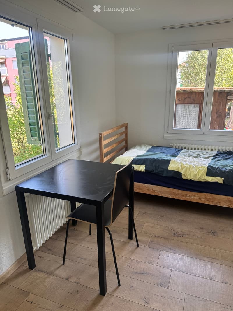 A furnished bedroom with a bed, a table, and a chair. The room has wooden flooring and large windows with a view of greenery outside.