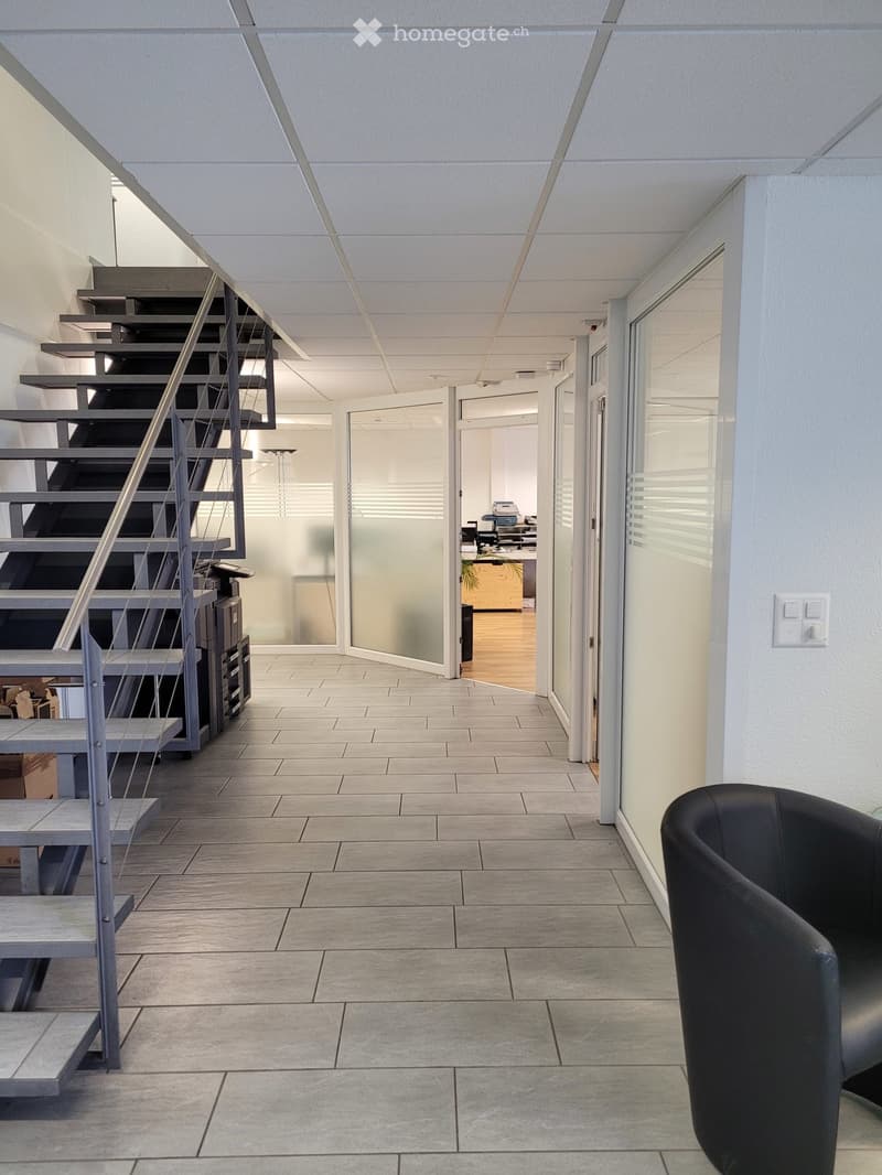 Modern hallway with tiled floor, staircase leading upwards, glass partitions, and a black leather chair in the foreground.