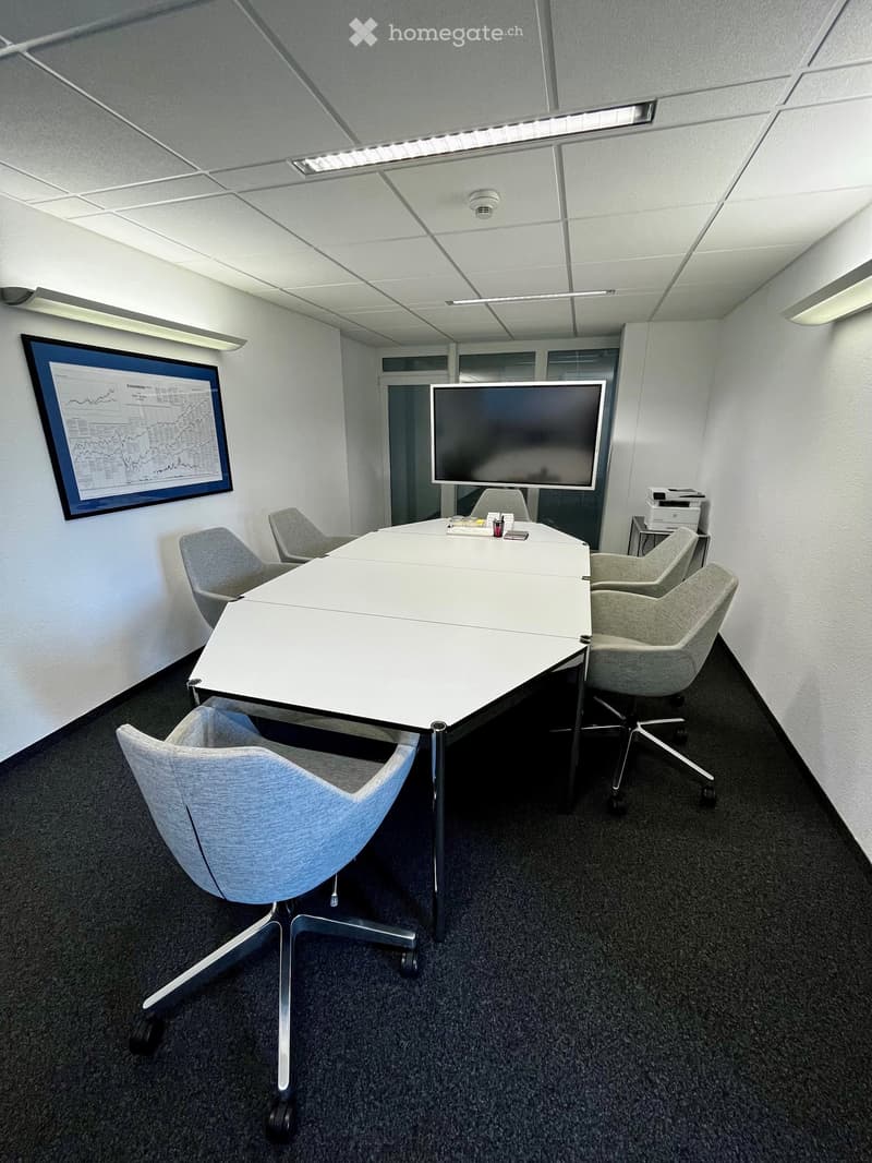 Modern office meeting room with a large white conference table, several grey chairs, a large screen, a printer, and a cup on the table.