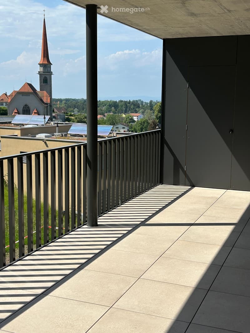Modern balcony with concrete floor, metal railing, and a view of a town with a church tower and surrounding landscape.