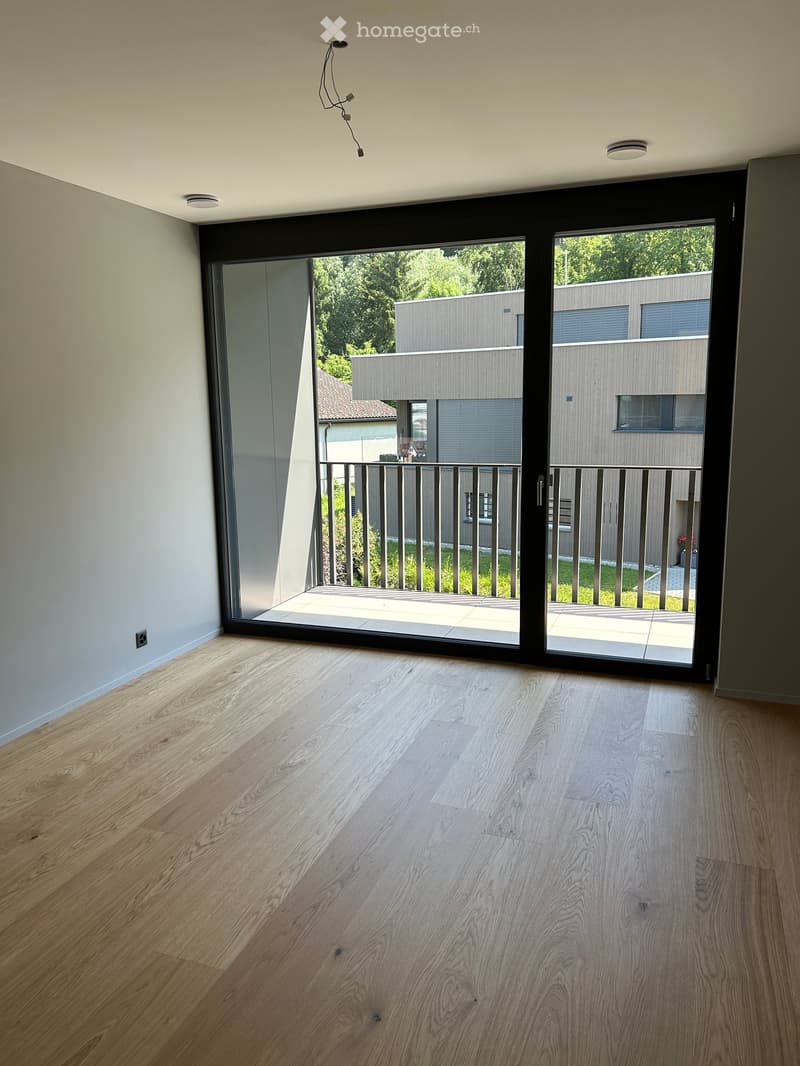 Empty room with light wooden flooring and large sliding glass doors leading to a balcony with a metal railing and greenery visible outside.