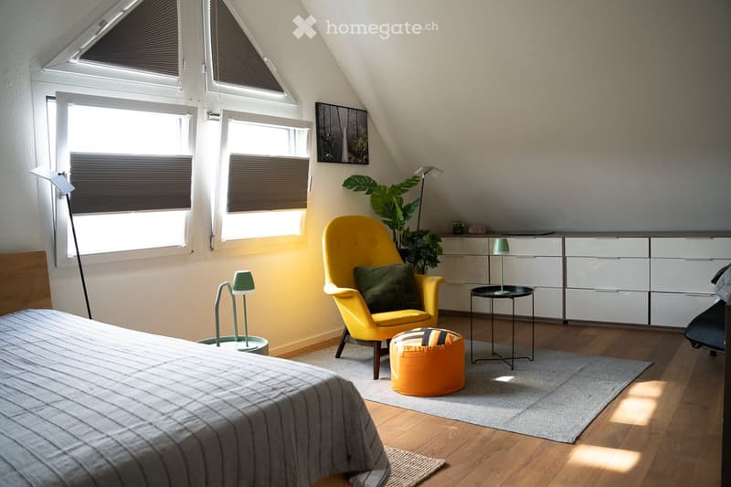 Bright attic bedroom with wooden floors, large windows, a yellow armchair, a round orange ottoman, a small black side table, and a modern bed.