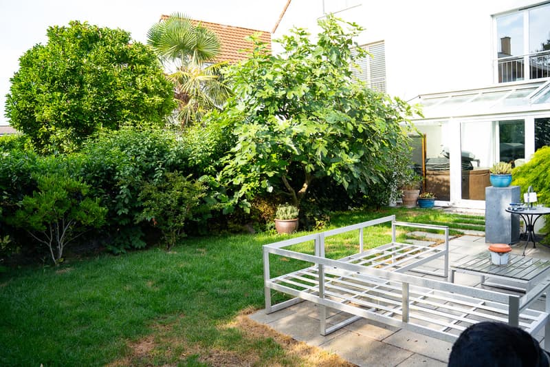 Well-maintained garden with green grass, various trees and shrubs, a modern metal bench, and a small patio area with a table and chairs.