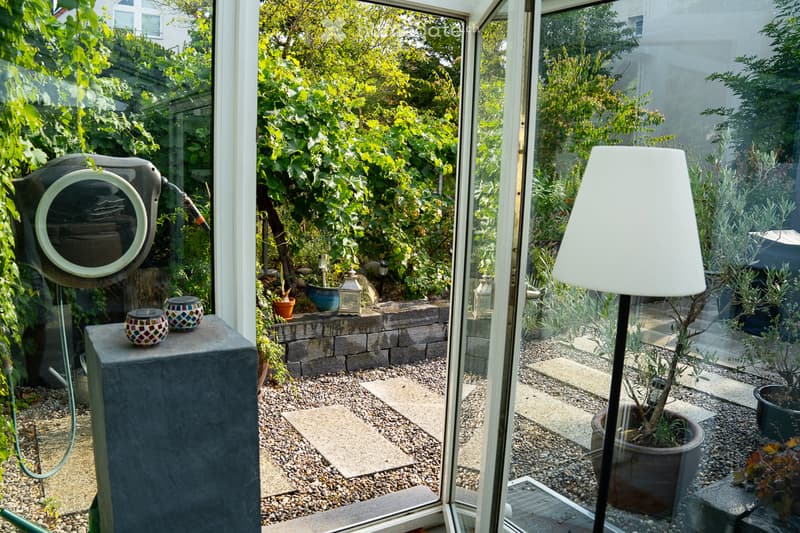 A well-maintained garden visible through large glass doors, featuring potted plants, gravel pathways, and decorative lanterns.