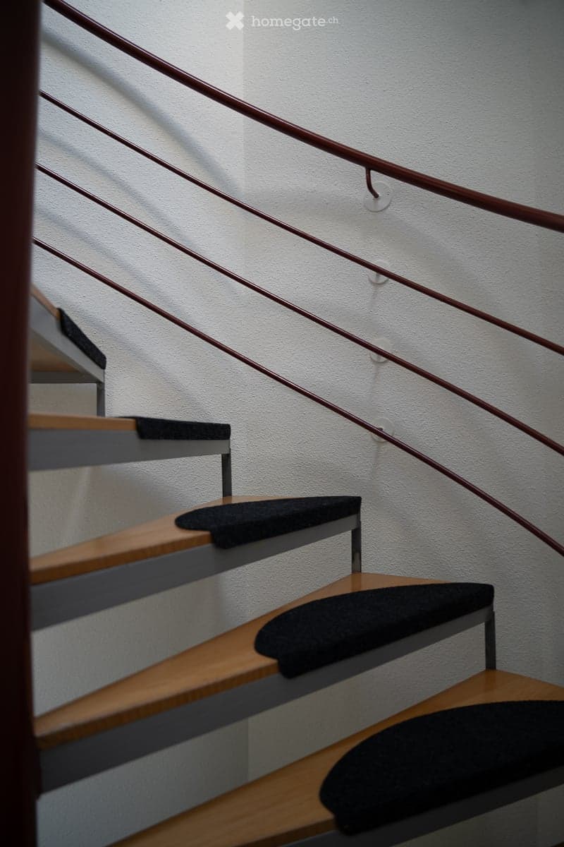Spiral staircase with wooden steps and dark-colored treads, featuring a metal railing and white walls.
