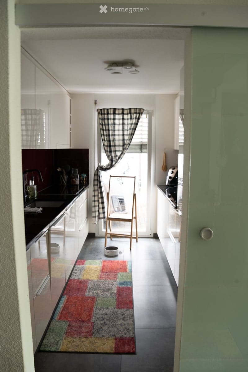 Modern kitchen with dark countertops, white cabinets, and a colorful rug. Features a sink, faucet, refrigerator, oven, microwave, and a small chair near the window.