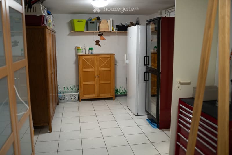 The kitchen features a white tiled floor, a wooden cabinet, a refrigerator, and shelves with various items. There is also a toolbox visible on the right side.