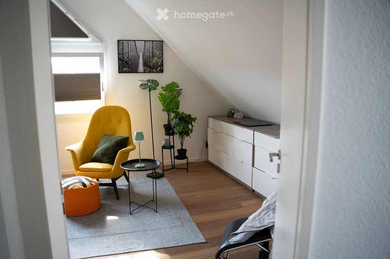 Bright, modern attic room with wooden flooring, large windows, a yellow armchair, ottoman, small round table, floor lamp, potted plants, and a white cabinet.