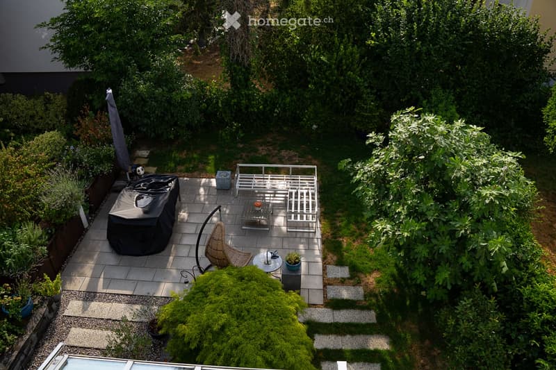 A well-maintained garden with a patio area featuring a wicker hanging chair, a small table, and a covered grill. The space is surrounded by lush greenery and shrubs, creating a serene outdoor setting.