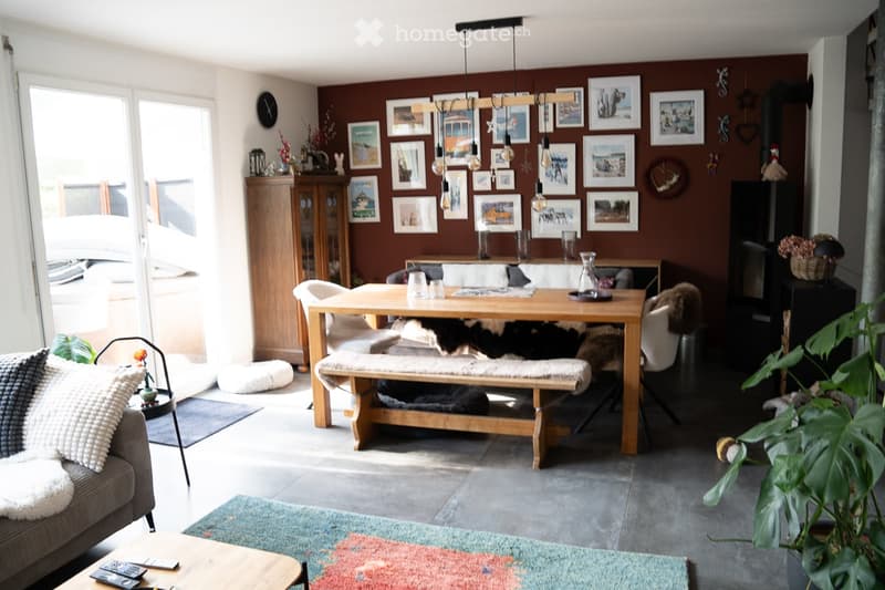 Cozy living and dining area with a grey sofa, coffee table, dining table with bench seating, fireplace, and various decorative items including picture frames and a plant.