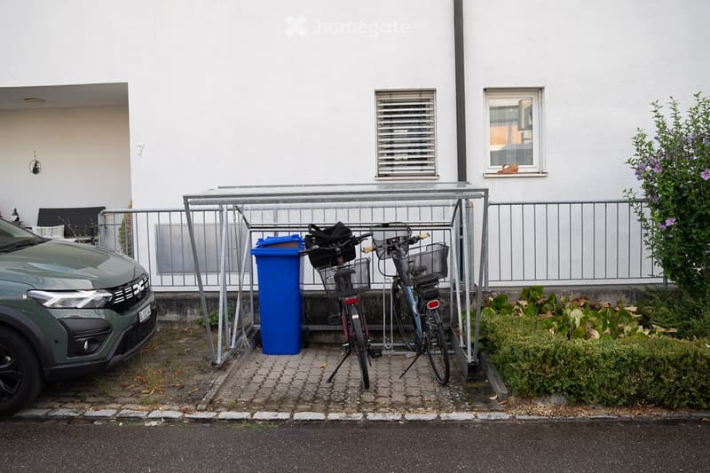 A modern building with a parking area featuring a car, two bicycles, and a blue recycling bin.
