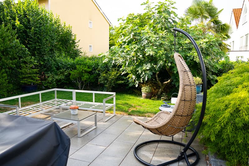 Garden area with a patio, white metal bench, glass coffee table, wicker hanging chair, and surrounding greenery.