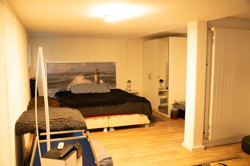 Cozy bedroom with wooden flooring, double bed with black duvet, nightstand with lamp, wardrobe, and a large picture of a lighthouse on the wall.