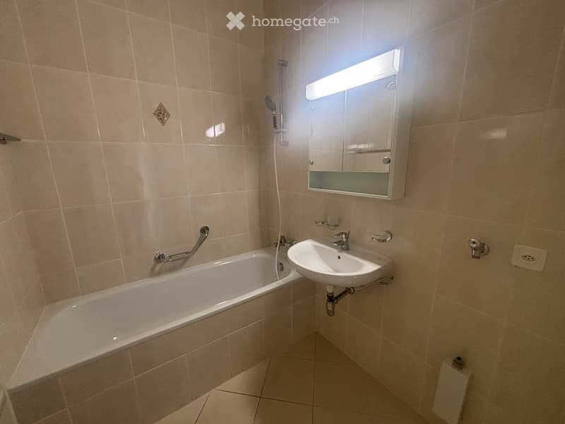 Bathroom with beige tiled walls and floor, featuring a bathtub, sink, mirror, and toilet.