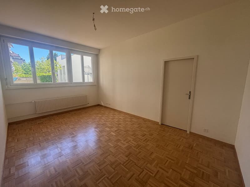 Empty room with wooden parquet flooring, white walls, large windows, and a radiator. No furniture or equipment present.