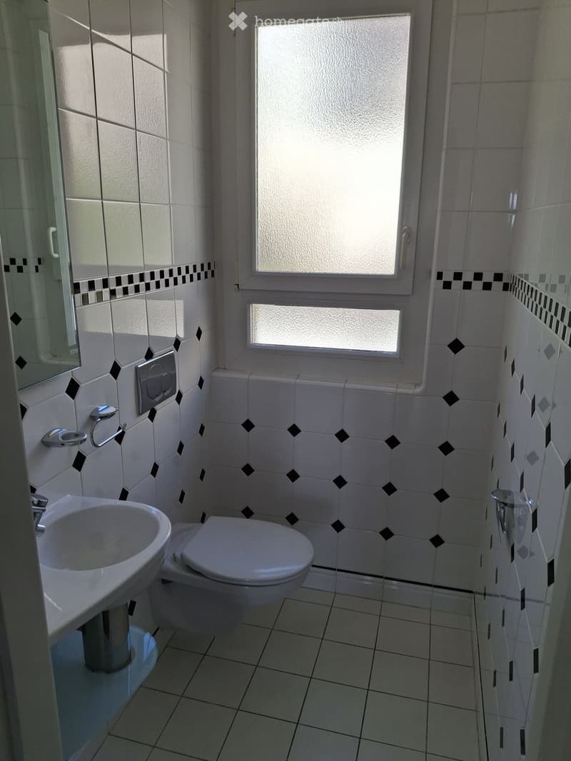 Modern bathroom with white tiles, black diamond pattern accents, a sink, toilet, and a mirror. Natural light from a frosted window illuminates the space.