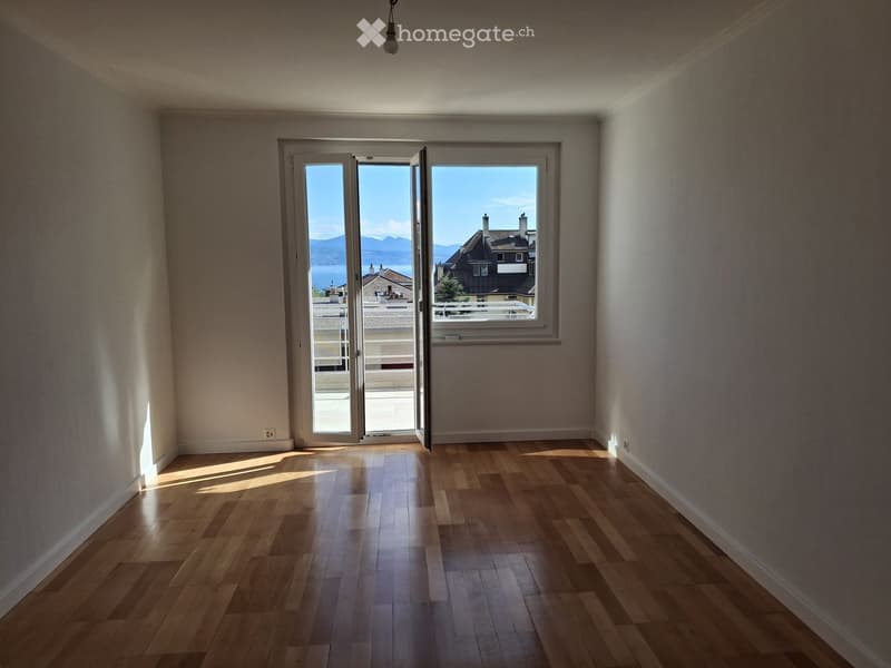 Empty room with wooden flooring, white walls, and large sliding glass doors leading to a balcony with a scenic view of mountains and a lake.