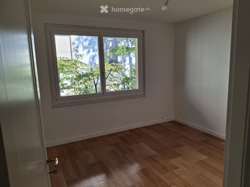 Empty room with light wooden flooring and white walls, featuring a large window with blinds and a view of greenery outside.