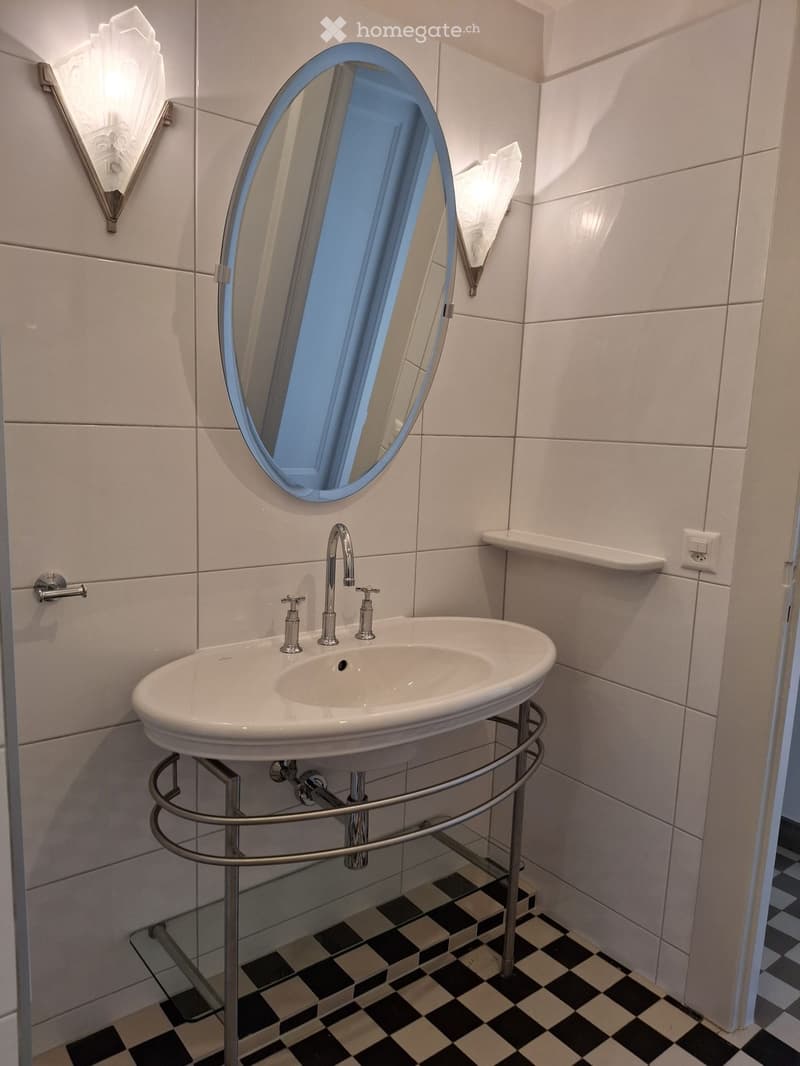 Modern bathroom with a white sink, chrome faucet, large oval mirror, and decorative wall-mounted light fixtures.
