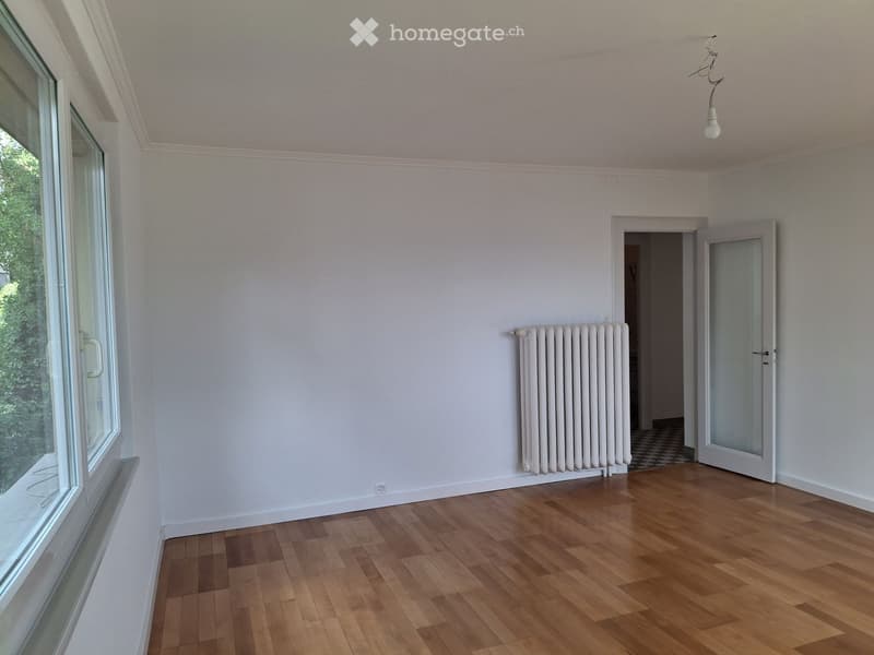 Empty room with wooden flooring, white walls, a radiator, and a large window with a view of greenery.
