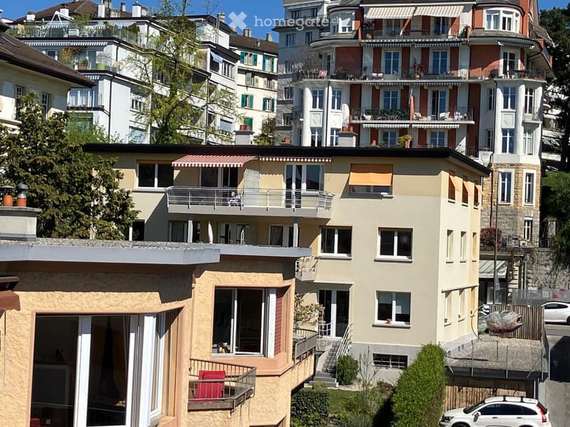 Multi-story residential building with balconies, large windows, and awnings, situated in a hillside neighborhood with neighboring houses and greenery.