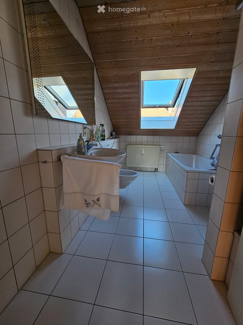 Modern bathroom with white tiled walls and floor, a sink, bathtub, toilet, and a towel hanging on the wall. The room features skylight windows and a radiator.