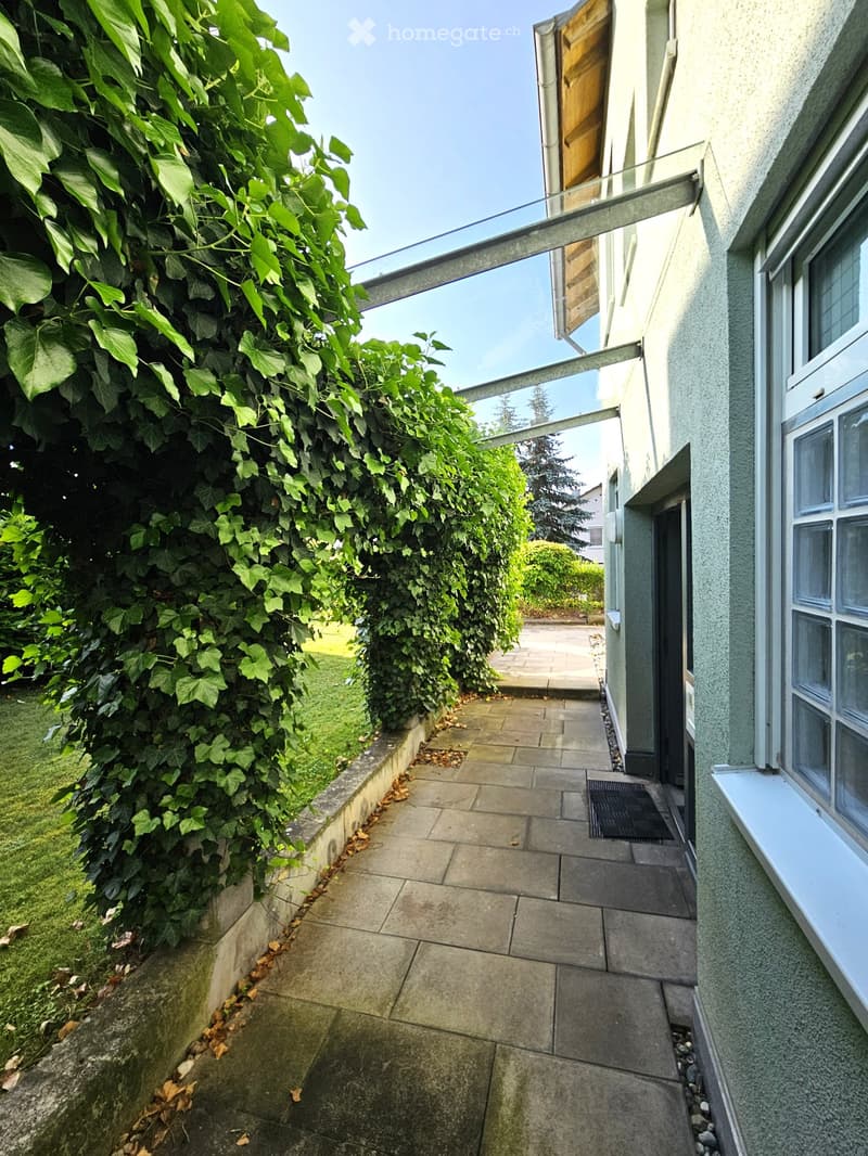 A narrow, paved balcony area with a glass roof structure, surrounded by lush green ivy and a grassy area.