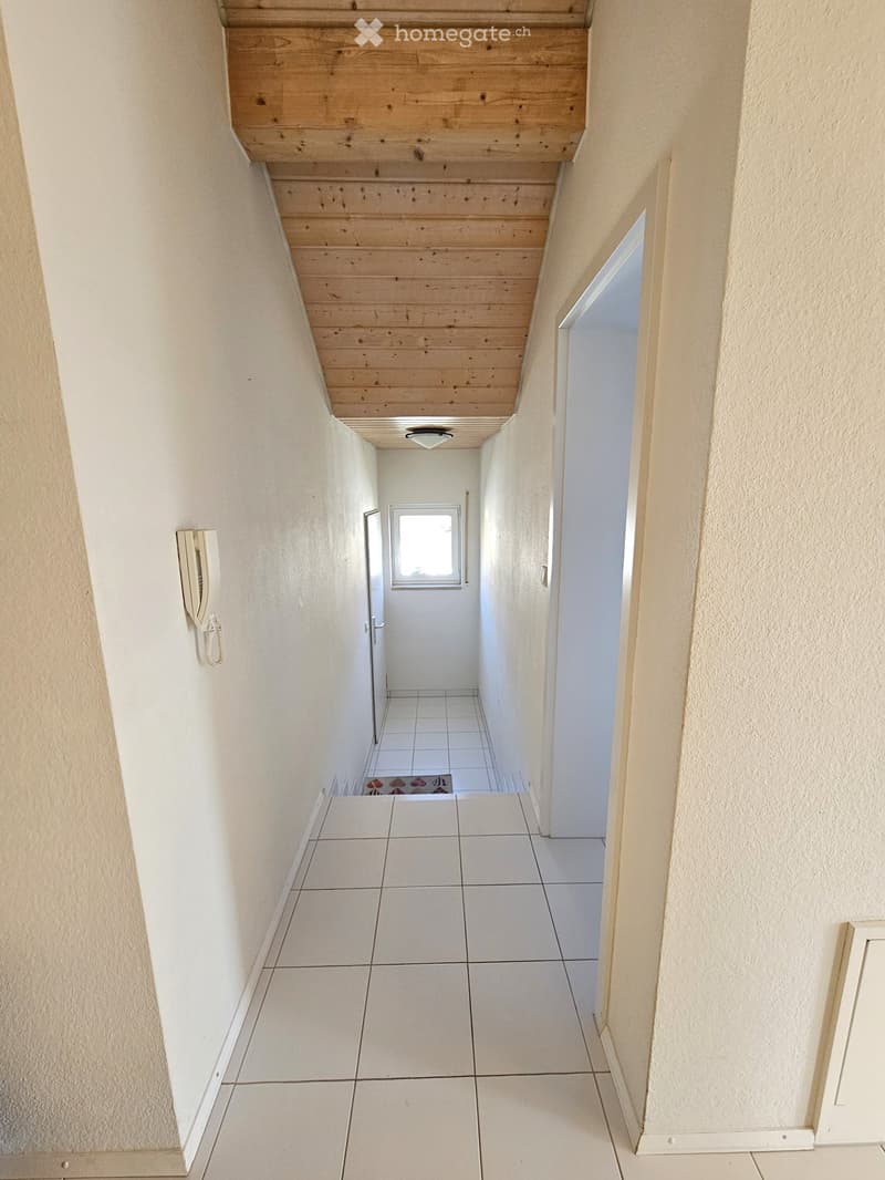 Empty hallway with white tiled flooring, light-colored walls, and a wooden slanted ceiling. A small window at the end provides natural light, and a wall-mounted phone is visible on the left side.