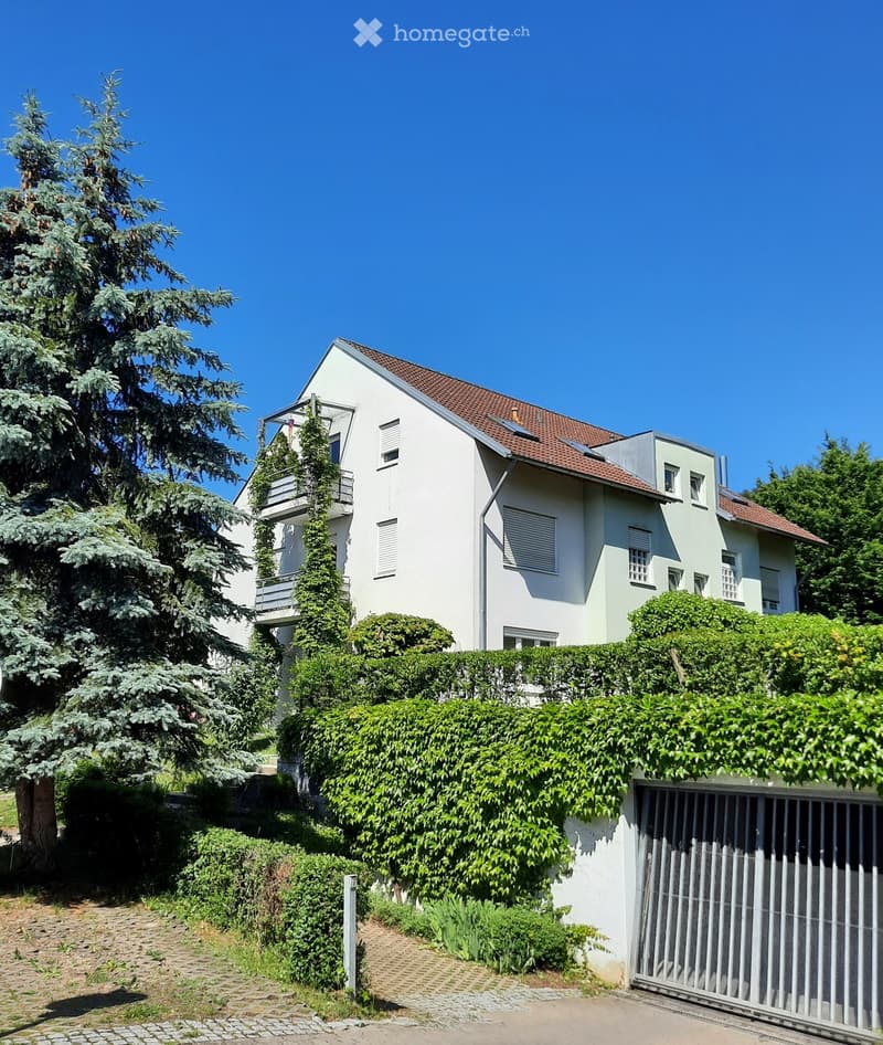 Two-story house with white and light green exterior, red-tiled roof, multiple windows, and a balcony on the upper level. Surrounded by greenery and a hedge.