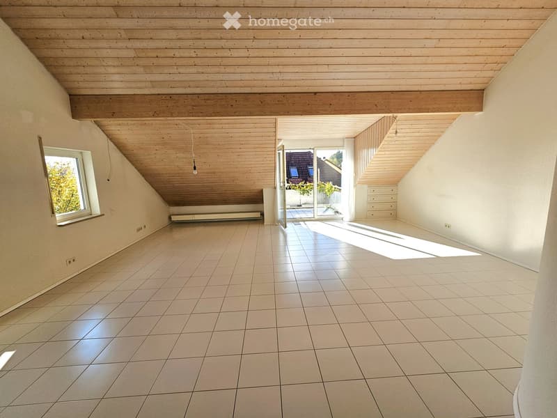 Empty attic room with wooden ceiling, tiled floor, large window, and sliding glass door leading to a balcony.
