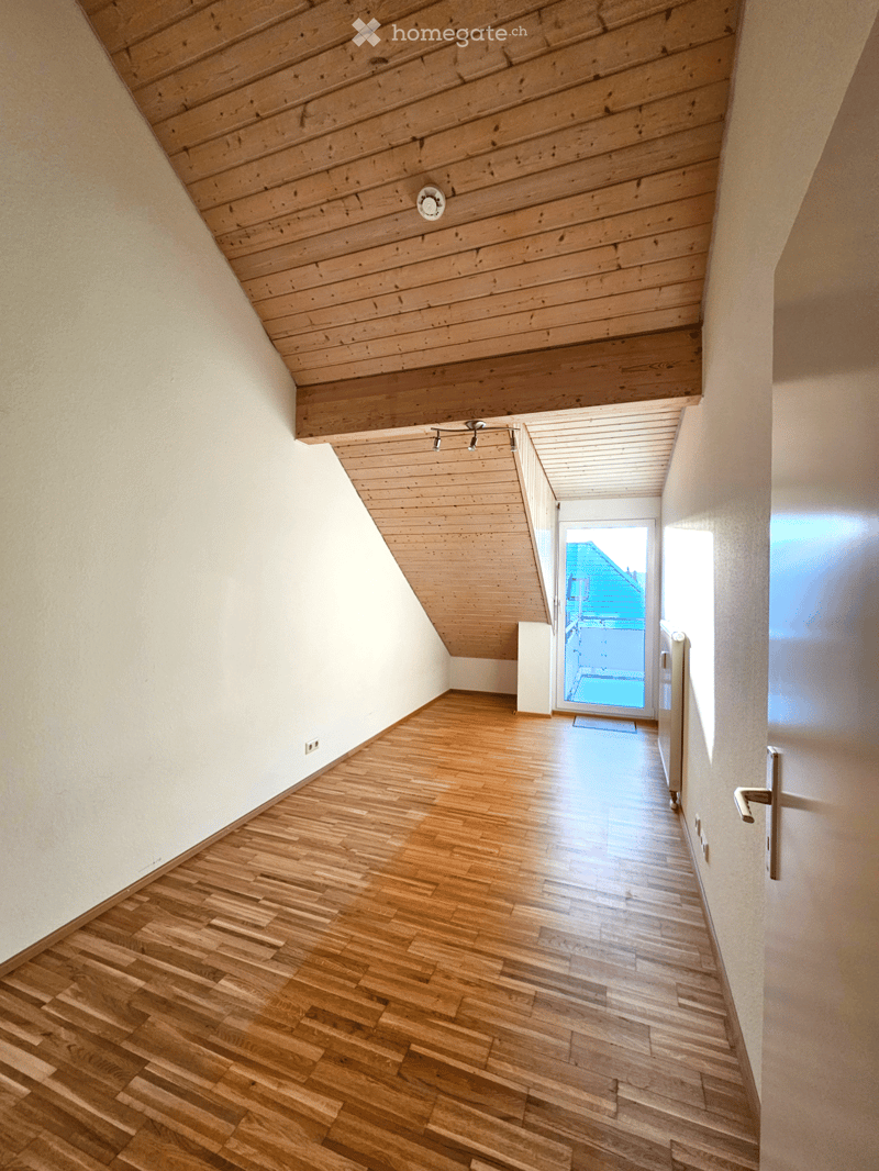 A narrow hallway with wooden flooring and white walls, featuring a slanted wooden ceiling and recessed lighting. An open doorway leads to a balcony area with a view of the outside.
