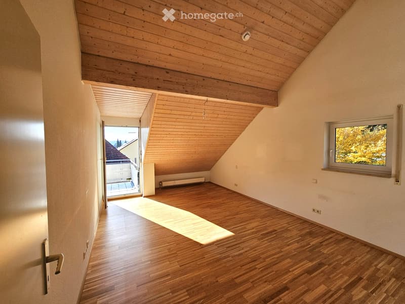 Bright, empty attic room with wooden floors and slanted wooden ceilings. Features a large window with a view of trees and an open doorway leading to a balcony area.