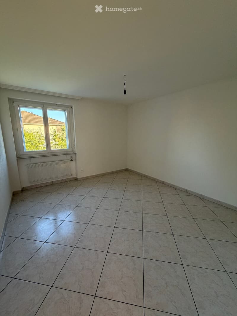 Empty room with tiled floor, light-colored walls, and a window with white frame. A radiator is visible below the window.