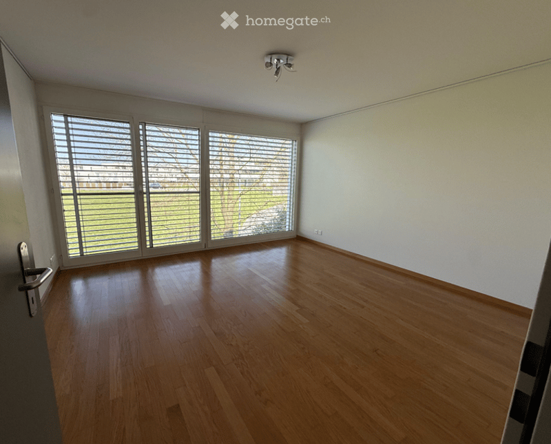 Empty room with wooden flooring, large windows with blinds, and a modern ceiling light fixture.