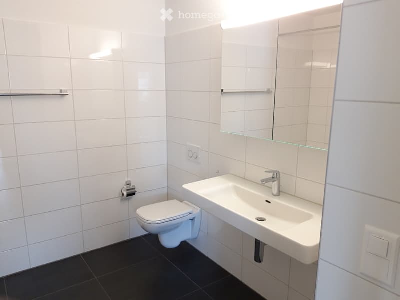 Modern bathroom with white tiles, a toilet, a sink with a chrome faucet, and a large mirror above the sink.