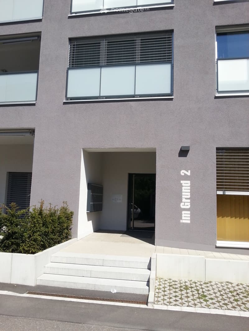 Modern two-story building with grey facade, large windows, and a raised ground floor entrance with steps.