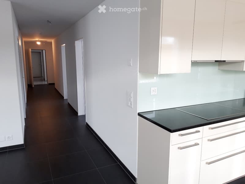 Empty hallway with white walls and dark tiled flooring, leading to a modern kitchen area with white cabinets and a black countertop.