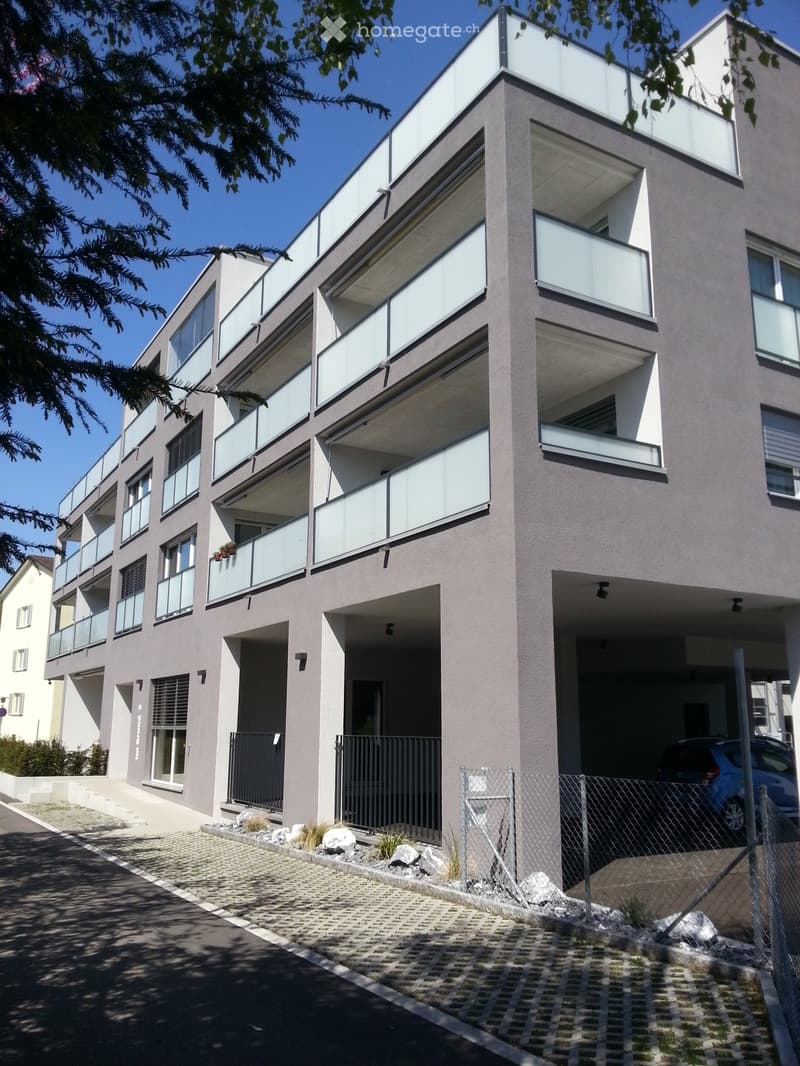 Modern multi-story residential building with balconies, large windows, and a paved entrance area.