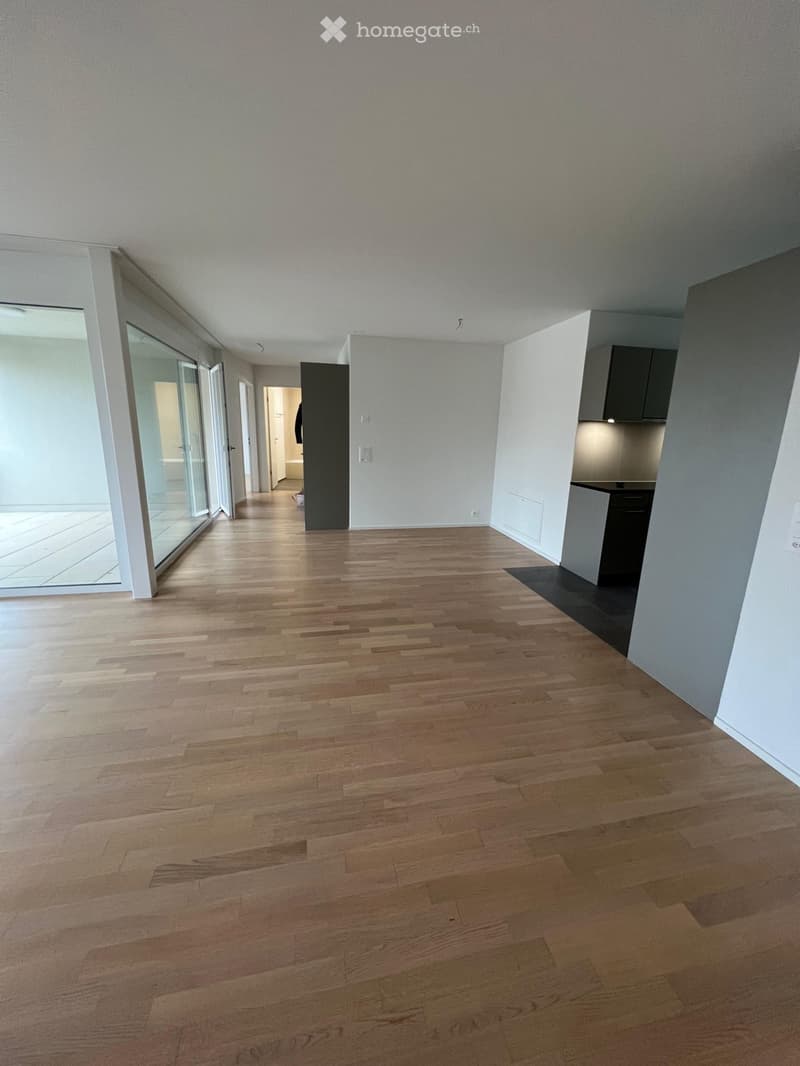 Empty hallway with wooden flooring, white walls, and a glass door leading to an outdoor area. A small kitchen area is visible on the right side.