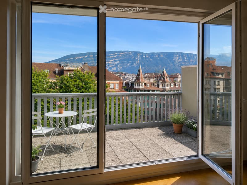 Balcony with a view of city rooftops and mountains, featuring a small table, two chairs, and potted plants.