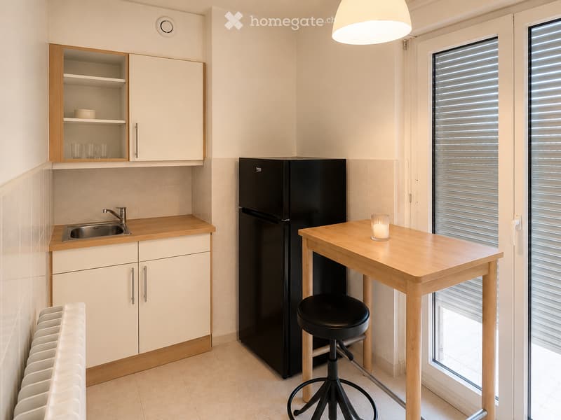 Modern kitchen with light wood cabinets, stainless steel sink, black refrigerator, wooden table with stool, and a candle on the table.